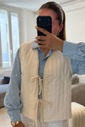 Gilet matelassé blanc pour femme avec liens à nouer et finition galon à pompons, porté sur une blouse bleu ciel et un pantalon blanc.