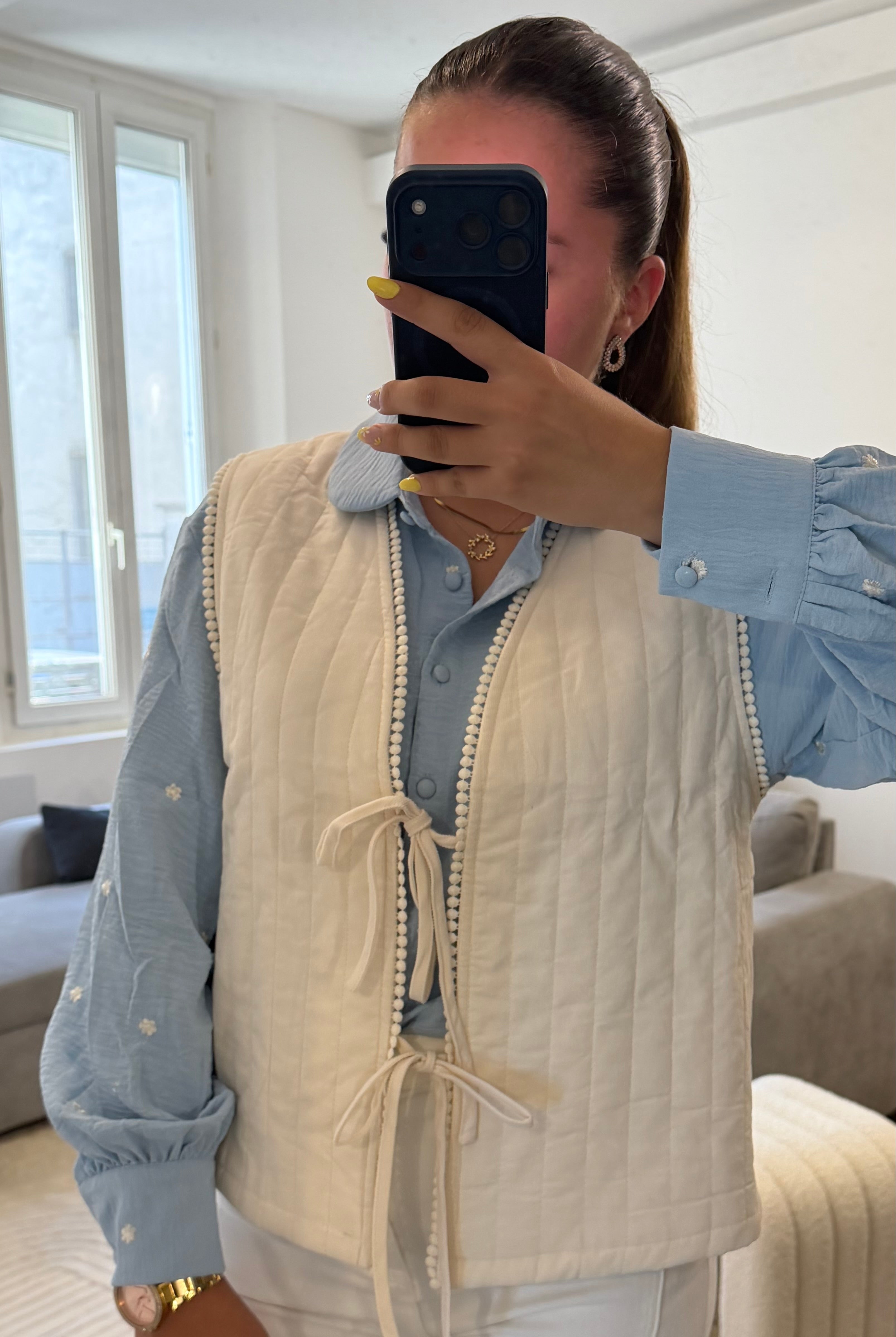 Gilet matelassé blanc pour femme avec liens à nouer et finition galon à pompons, porté sur une blouse bleu ciel et un pantalon blanc.