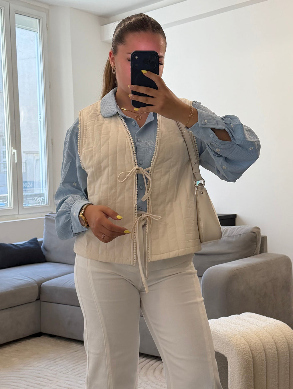 Gilet matelassé blanc pour femme avec liens à nouer et finition galon à pompons, porté sur une blouse bleu ciel et un pantalon blanc.