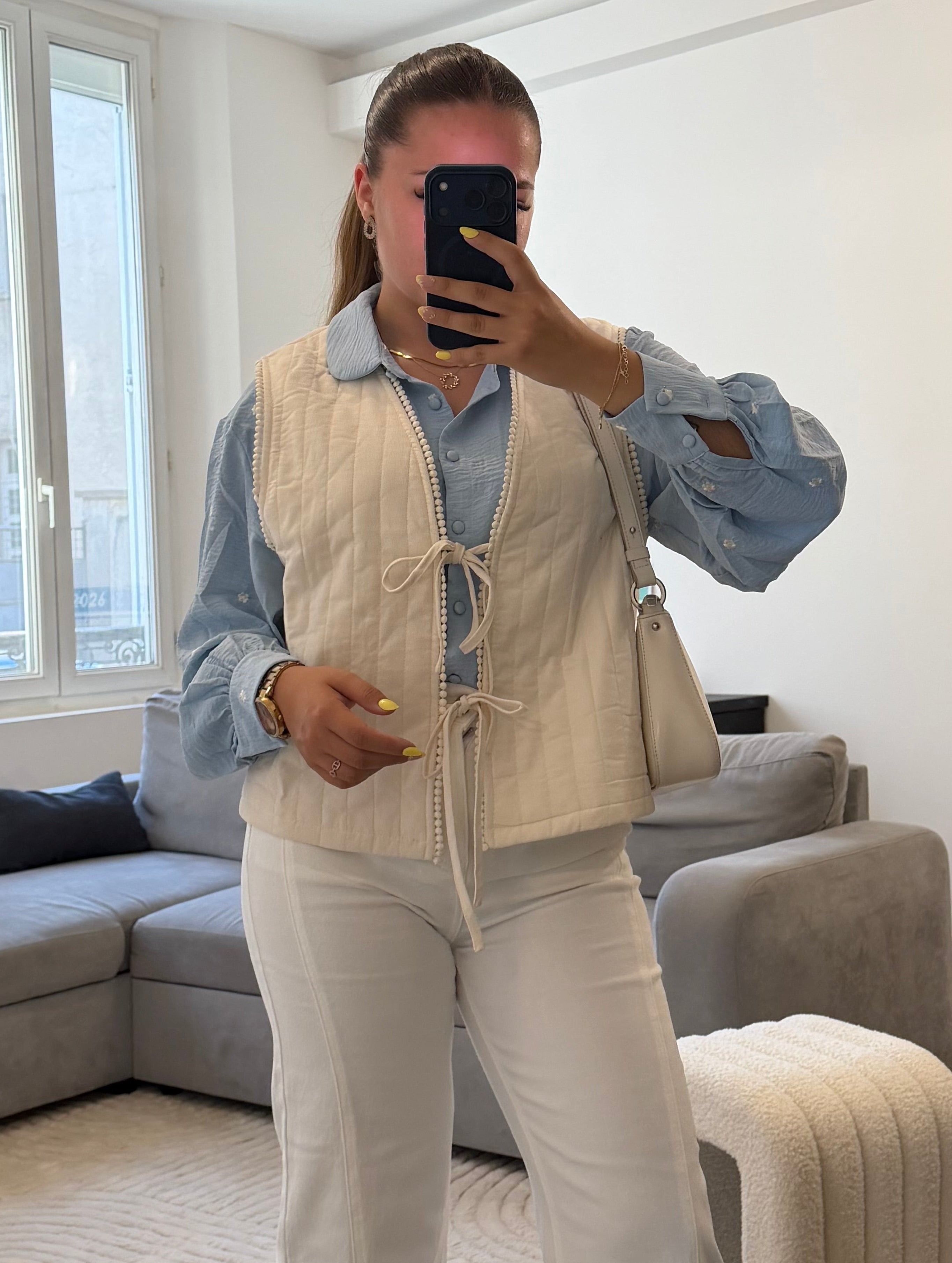 Gilet matelassé blanc pour femme avec liens à nouer et finition galon à pompons, porté sur une blouse bleu ciel et un pantalon blanc.
